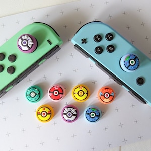 May include: Eight colourful round joystick caps with different Pokemon designs. The caps are on a white background with a plus sign pattern. Two Nintendo Switch controllers are in the background, one green and one blue.