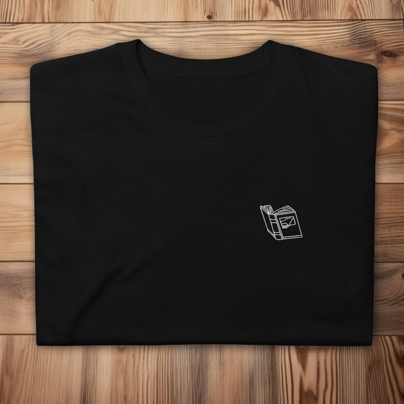 May include: Black t-shirt with a white outline of an open book embroidered on the front.