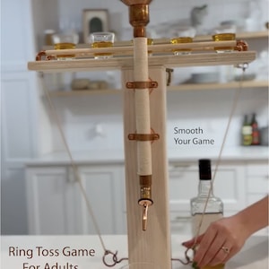 May include: A wooden ring toss game with a copper filtration system for spirits and wine. The game features a copper funnel, a wooden base, and a copper tap. The text "COLLIDE REFINING COLUMN" is printed on the top of the game. The text "Smooth Your Game" is printed on the side of the game. The text "Ring Toss Game For Adults" is printed on the base of the game.