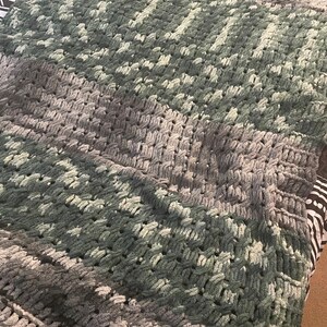 May include: A soft, crocheted throw blanket in shades of green and gray. The blanket has a textured, woven pattern.