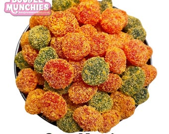 May include: A bowl filled with colorful, round candies. The candies are coated in a granular substance and come in shades of orange, green, and red. The text "Sour Mango" is displayed at the bottom of the image, with the brand logo "Boujee Munchies" at the top.