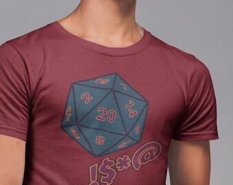 Puede incluir: Una camiseta de manga corta color granate con un gráfico de un dado de veinte caras azul oscuro con números rojos, incluyendo un 20 prominente. Debajo del dado, el texto !$%*@ está impreso en una fuente roja estilizada con borde oscuro.