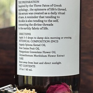 May include: A brown glass bottle with a white label featuring text about a serum inspired by Greek mythology. The label lists ingredients and directions. A dried pink rose and a dried flower are in the foreground. The bottle contains 2 fl oz / 60 mL.