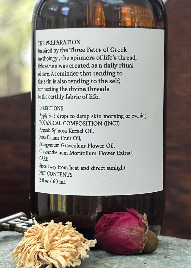 May include: A brown glass bottle with a white label featuring text about a serum inspired by Greek mythology. The label lists ingredients and directions. A dried pink rose and a dried flower are in the foreground. The bottle contains 2 fl oz / 60 mL.