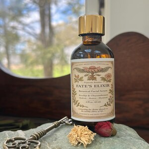 May include: A brown glass bottle of "FATE'S ELIXIR" botanical facial serum with a gold dropper. The label reads "Roschip & Chrysanthemum" and "2 fl oz - 60 ml". A decorative key and dried flowers rest on a stone.