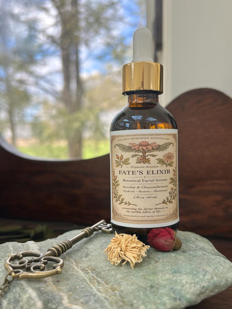 May include: A brown glass bottle of "FATE'S ELIXIR" botanical facial serum with a gold dropper. The label reads "Roschip & Chrysanthemum" and "2 fl oz - 60 ml". A decorative key and dried flowers rest on a stone.