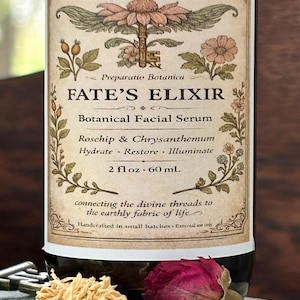 May include: A brown glass bottle with a label that reads "FATE'S ELIXIR Botanical Facial Serum." The label features botanical illustrations and text. The bottle contains 2 fl oz, which is equivalent to 60 mL. Dried flowers and a key are in the foreground.