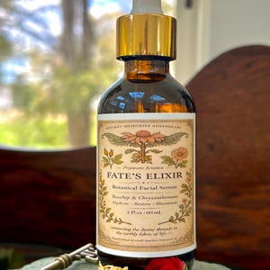 Puede incluir: Una botella de vidrio marrón de suero facial botánico Fate's Elixir con un cuentagotas dorado. La etiqueta dice "Fate's Elixir" y enumera los ingredientes. La botella es de 60 ml. Una llave y flores secas están en primer plano.