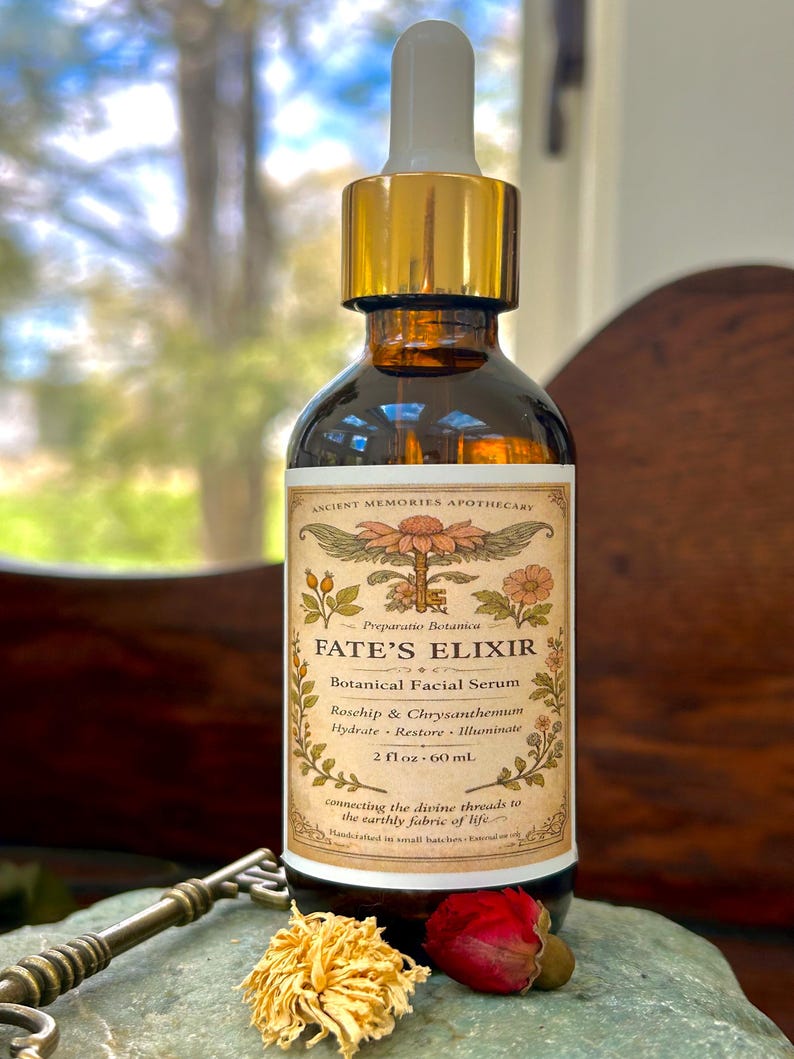 May include: A brown glass bottle of Fate's Elixir botanical facial serum with a gold dropper. The label reads "Fate's Elixir" and lists ingredients. The bottle is 2 fl oz (60 ml). A key and dried flowers are in the foreground.