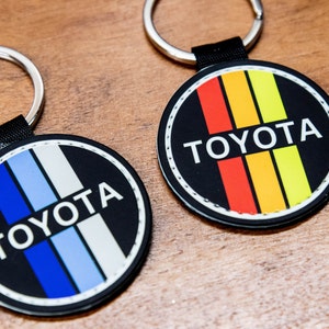 May include: Two black round keychains with a white stitched border. Each keychain has a black background with a white "TOYOTA" logo and vertical colored stripes. One keychain has blue and white stripes, and the other has red, orange, and yellow stripes.