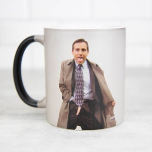 May include: A white ceramic mug with a black handle features a color image of a man in a suit and coat sticking his tongue out. The man is wearing a patterned tie.