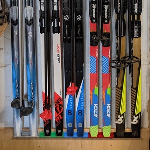 May include: A collection of nine pairs of skis and poles hanging on a wall. The skis are various colors, including blue, red, yellow, and black. The poles are black and have a variety of brands, including Salomon, Rossignol, and Fischer.