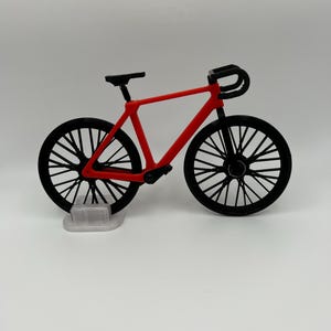 May include: A red toy bicycle with black wheels and handlebars. The bicycle is on a white stand.