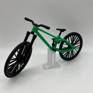 May include: A green and black miniature mountain bike on a clear plastic stand. The bike has a black frame, black wheels, and a black handlebar.
