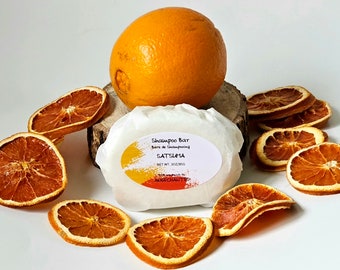 May include: A white shampoo bar wrapped in paper with the text "Shampoo Bar, Barre de Shampoing, SATSUMA, NET WT. 3OZ/85G, ALCHECHARM" printed on it. The bar is surrounded by dried orange slices.