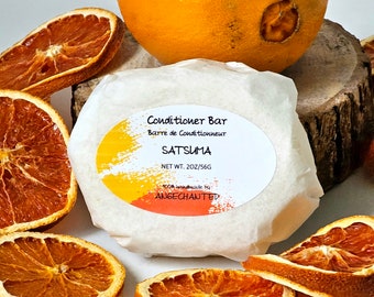 May include: A white conditioner bar wrapped in parchment paper with a label that reads "Conditioner Bar, Barre de Conditionneur, SATSUMA, NET WT. 2OZ/56G, 100% handmade by ANGECHANTED". The bar is surrounded by dried orange slices.