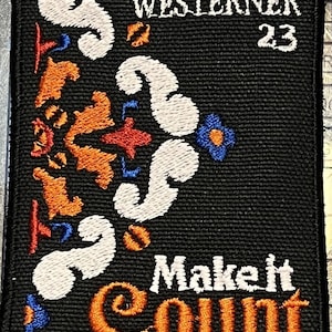 Peut inclure: Écusson brodé noir avec les mots "WESTERNER 23" en haut. Le patch présente des motifs floraux blancs, orange, rouges et bleus et la phrase "Make it Count" en orange.