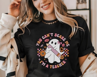 May include: Black t-shirt featuring a white ghost holding a pink pencil. The ghost has a pink bow and the text "You can't scare me I'm a teacher" in pink and white lettering.
