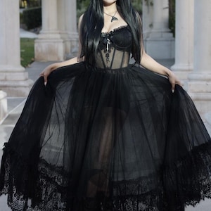 May include: A woman in a black sheer gown with a lace trim. The gown has a corset top with a keyhole detail and a black lace overlay. She is wearing a silver necklace with a pendant. The woman is standing in front of a white column.