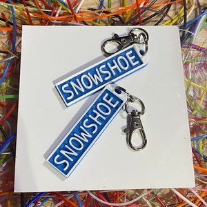 May include: Two blue keyrings with the word "SNOWSHOE" printed in white letters. The keyrings are attached to silver key rings.