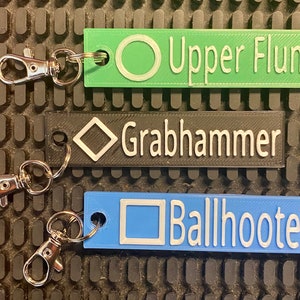 May include: Three keychains with different coloured plastic tags. The green tag says "Upper Flume" with a circle symbol. The black tag says "Grabhammer" with a diamond symbol. The blue tag says "Ballhooter" with a square symbol.