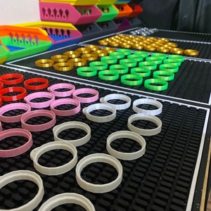 May include: A black mat with rows of colourful plastic rings. The rings are arranged in rows of different colours, including white, pink, red, green, gold, and black.