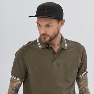 May include: A black 5-panel cap with a red embroidered "YOU DIED" text on the front. The cap is worn by a person wearing a green polo shirt with white trim.