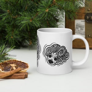 May include: White ceramic mug with a black and white abstract design. The design features a stylized face with swirling patterns. The mug has a curved handle. The background includes greenery and decorative items.