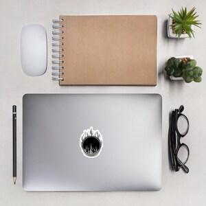 May include: A black and white sticker with a circular design and a flame-like border. The sticker is on a grey laptop, with a white mouse, a brown notebook, a pencil, glasses, and small potted plants around it.