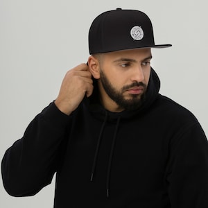 Puede incluir: Una gorra de béisbol negra ajustada con un logotipo bordado en blanco y negro. El logotipo presenta un círculo con un rayo y una S estilizada en su interior.