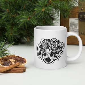 May include: White ceramic mug with a black tribal face design and swirling hair. The mug has a curved handle. The background includes greenery and decorative items.