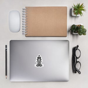 May include: A black and white sticker depicting a detailed illustration of a figure with a tall, pointed hat. The sticker is on a laptop, with a mouse, notebook, pencil, glasses, and small potted plants arranged around it.