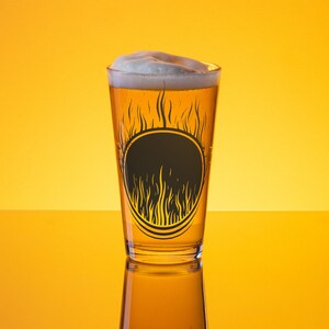 May include: A pint glass filled with beer and a foamy head. The glass has a black circular design with flame-like details. The background is a gradient of yellow and orange.