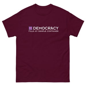 May include: A maroon t-shirt with the word "DEMOCRACY" in white capital letters, above the phrase "FILLS MY SAMPLE CONTAINER." The word "DEMOCRACY" is accompanied by a small, multi-colored square graphic. The shirt is made of a soft material.