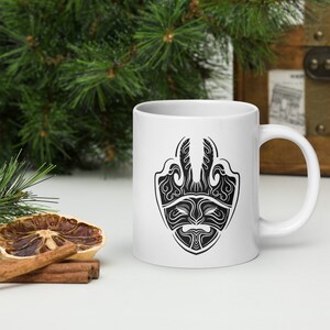 May include: White ceramic mug with a black tribal mask design. The mask features detailed symmetrical patterns. The mug has a standard handle and is suitable for hot or cold drinks.