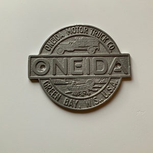 May include: A silver metal emblem with the text "Oneida Motor Truck Co." and "Oneida" in a larger font. The emblem also features a small image of a truck and the text "Green Bay, Wis. U.S.A."