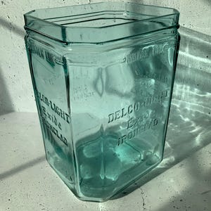 Peut inclure: Un bocal en verre turquoise hexagonal vintage. Le bocal porte les inscriptions "WATER LINE", "DELCO-LIGHT Exide IRONCLAD" en relief sur ses côtés. Le bocal est vide et posé sur une surface blanche, avec des ombres.
