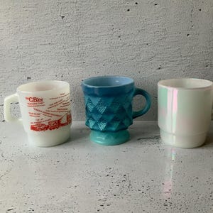 Könnte beinhalten: Drei weiße Keramikbecher mit unterschiedlichen Designs. Ein Becher hat ein rot-weißes Design mit Text, ein anderer ein blaues und grünes geometrisches Muster und der dritte hat eine Perlmutt-Oberfläche.