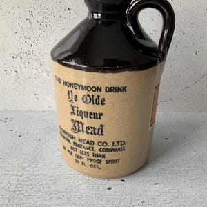 Op de afbeelding: Een bruine keramische kan met een kurkstop en een handvat. De kan is gelabeld "The Honeymoon Drink Le Olde Liqueur Mead Cornish Mead Co. Ltd.  Nolty, Penzance, Cornwall Not Less Than 20 Per Cent Proof Spirit 20 Fl. Ozs."