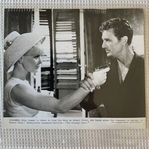 May include: A black and white photo of a woman in a hat pointing a gun at a man. The text on the photo reads "DISARMED: Elke Sommer is about to lose the drop on Robert Stack, who knows where the treasure is buried, in Warner Bros. Technicolor suspense-thriller, "The Corrupt Ones."