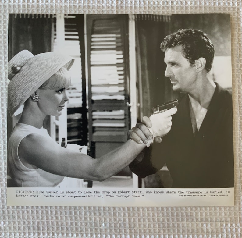 May include: A black and white photo of a woman in a hat pointing a gun at a man. The text on the photo reads "DISARMED: Elke Sommer is about to lose the drop on Robert Stack, who knows where the treasure is buried, in Warner Bros. Technicolor suspense-thriller, "The Corrupt Ones."