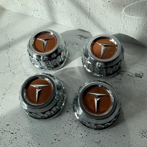 May include: Four vintage chrome hubcaps with a brown center and a silver T-shaped emblem. The hubcaps have a ribbed, textured design around the edges. The hubcaps are arranged on a light gray surface.