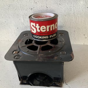 Puede incluir: Una lata de combustible de cocina Sterno vintage en rojo y blanco, encima de una estufa de metal negro. La lata es cilíndrica con el logotipo de Sterno e información del producto. La estufa tiene una base cuadrada con un quemador circular.