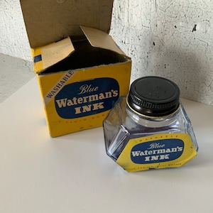 Pode incluir: Uma garrafa de vidro de tinta azul com uma tampa preta. A garrafa é etiquetada com "Blue Waterman's Ink" e "Washable". A garrafa está sobre uma superfície branca. A garrafa está em uma caixa amarela com o mesmo rótulo.
