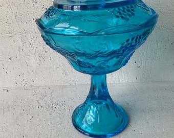 Puede incluir: Un plat de dulces de vidrio azul con tapa y base de pedestal. El plato está decorado con un patrón de vid.
