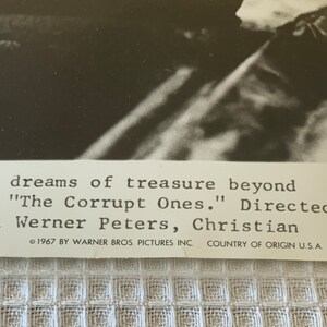 May include: Black and white photo of a movie poster for the film "The Corrupt Ones." The poster features a close-up of a man's face. The text on the poster reads "their dreams of treasure beyond" and "Directed by Kwan, Werner Peters, Christian."