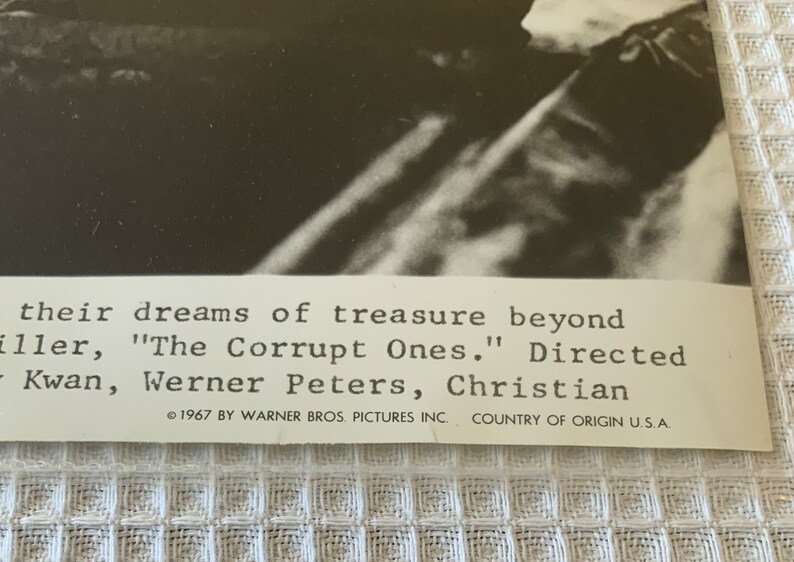 May include: Black and white photo of a movie poster for the film "The Corrupt Ones." The poster features a close-up of a man's face. The text on the poster reads "their dreams of treasure beyond" and "Directed by Kwan, Werner Peters, Christian."