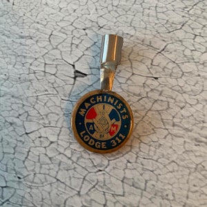 Puede incluir: Un pin de metal dorado con un logotipo azul, rojo y blanco que dice "Machinists Lodge 311 A. of M.". El pin está unido a un porta lápices de metal plateado.