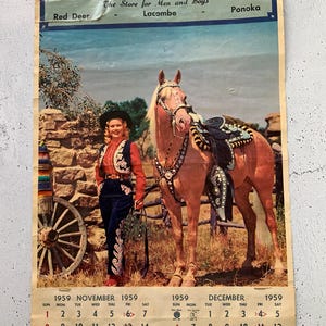 Op de afbeelding: Een vintage kalender met een vrouw in een cowboy-outfit die een paard met een zadel vasthoudt. De kalender is voor Jack's Men's Wear, een winkel voor mannen en jongens in Red Deer, Lacombe en Ponoka. De kalender toont de maanden november en december 1959.