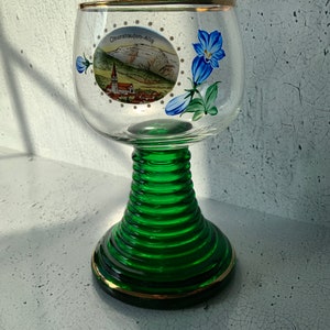 May include: A green glass wine glass with a gold rim and a painted scene of a village with the text "Oberstaufen-Allg" on the side. The glass has a spiral stem and a round base.